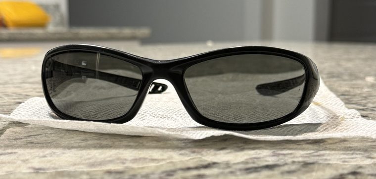 The original pair: glossy black wraparound sunglasses with smoke lenses, sitting on a paper towel on a kitchen counter